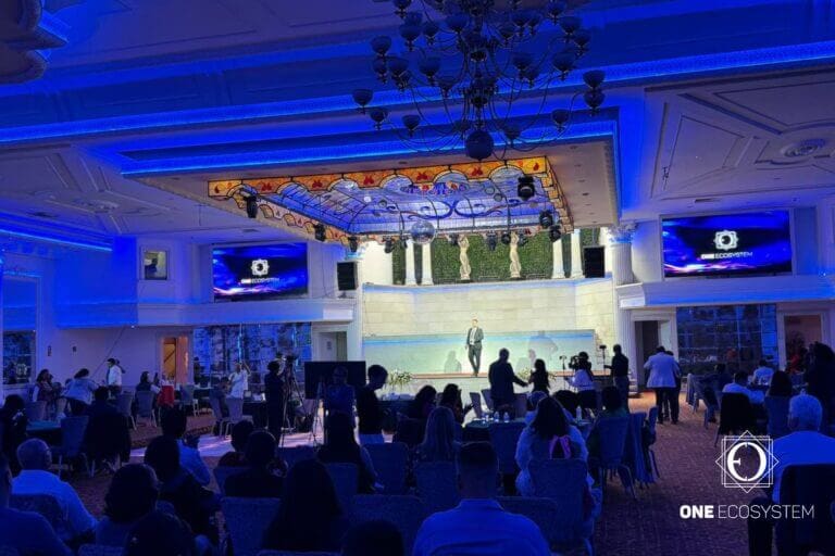 Nella sala conferenze, le persone sono sedute di fronte al palco dove si trova l'oratore. La sala è illuminata da luci blu, con grandi lampadari e decorazioni a soffitto. Mentre l'oratore elabora la tecnologia stand-alone, lo schermo visualizza "ONE ECOSYSTEM".