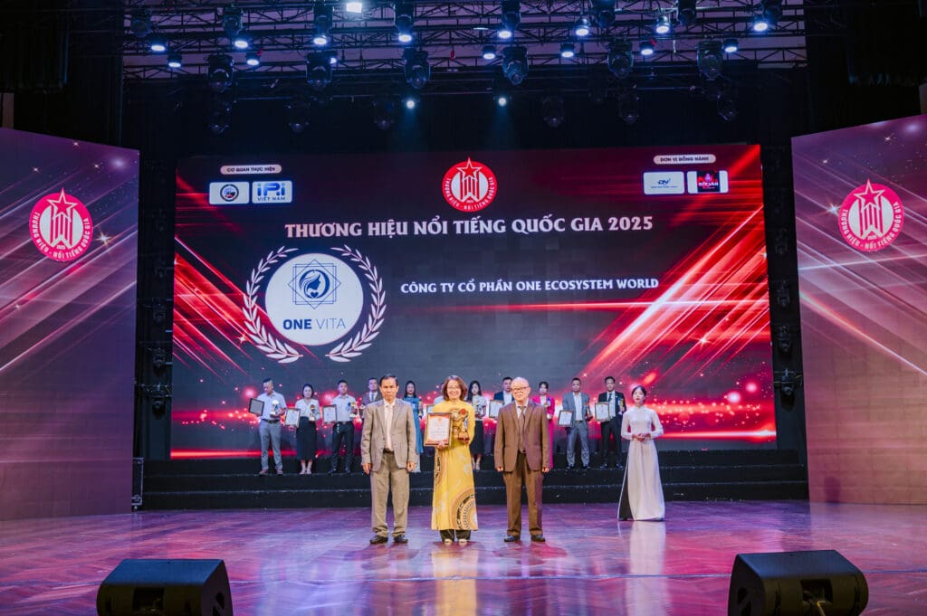 Persone sul palco durante la cerimonia di premiazione. Una donna in abito giallo tiene un certificato incorniciato, affiancata da due uomini in abito elegante. Sullo sfondo, il logo OES e la scritta del marchio nazionale certificato del Vietnam sono proiettati su un grande schermo.