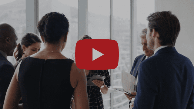 In un ufficio luminoso, persone vestite in modo professionale tengono in mano documenti e tablet e conversano su fattori che cambiano le carte in tavola. Un grande pulsante di riproduzione si sovrappone all'immagine, indicando il video.