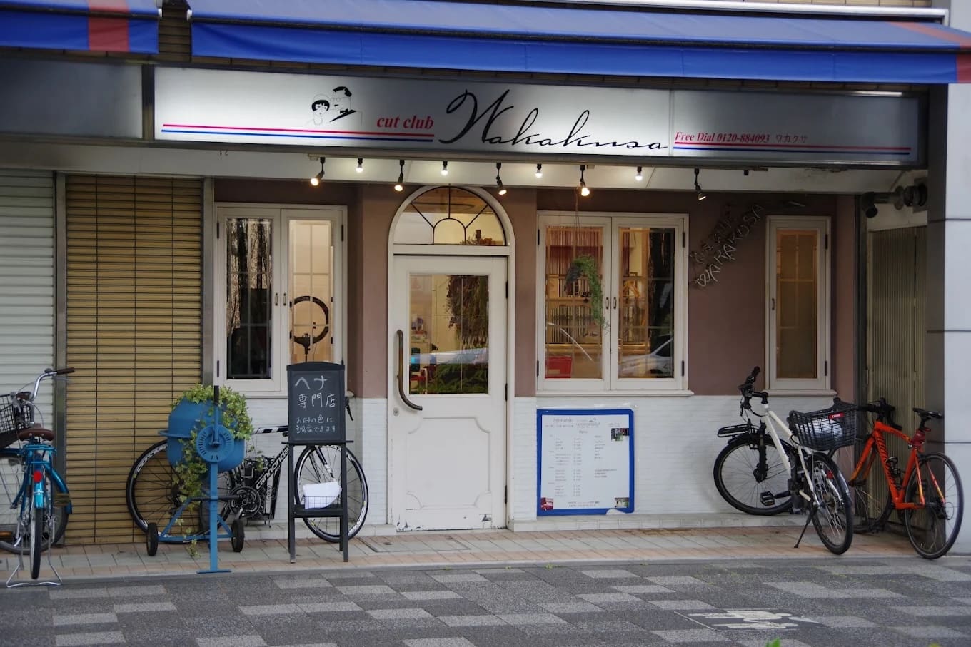 Il negozio ha un cartello che dice "cut club Wakana" in cima a una doppia porta bianca e offre servizi di scorciatoie e henné. Stretti tra le finestre e il parcheggio per biciclette, ci sono rastrelliere blu per biciclette, piante d'appartamento e menu e insegne vicino all'ingresso.