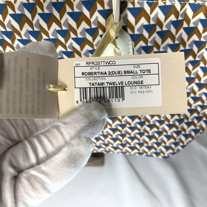 La mano guantata sorregge il cartellino del prodotto "ROBERTINA 2 (DUE) SMALL TOTE" sopra il tessuto con motivo tatami con motivo geometrico nei colori blu, marrone e bianco, che è la cifra stilistica della collezione di piccole tote di ROBERTA PIERI.