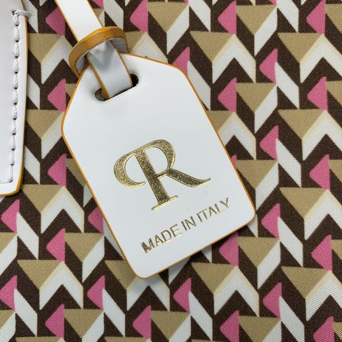 Un primo piano di un'etichetta bagaglio bianca di Roberta Pieri con la scritta "R" in oro e la scritta "MADE IN ITALY" attaccata a un mini borsone con motivi geometrici marroni, beige e rosa.