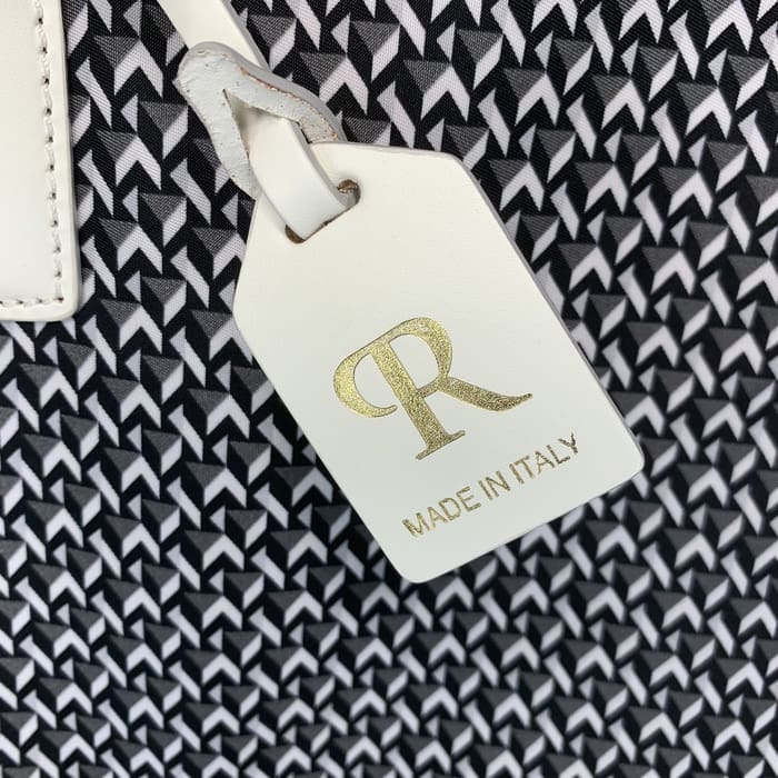 Una targhetta ROBERTA PIERI in pelle bianca con le lettere "R" in oro e la scritta "MADE IN ITALY" pende dallo sfondo di un tessuto geometrico bianco e nero.