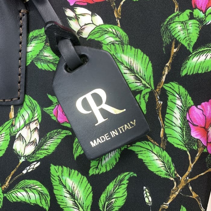 L'etichetta nera ROBERTA PIERI con la "R" dorata e il testo italiano è sospesa a un tessuto floreale con foglie verdi e fiori rosa, completando il mini borsone con un'eleganza italiana senza tempo.