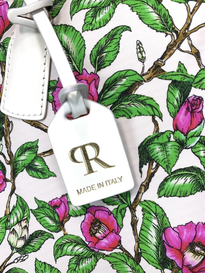Primo piano del mini borsone ROBERTA PIERI. peduncoli con foglie verdi e fiori rosa; L'etichetta bianca attaccata alla borsa a 2 vie presenta un logo "R" dorato e la scritta "MADE IN ITALY".